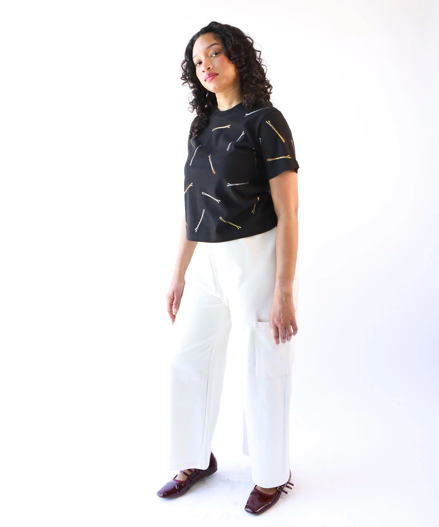 Woman wearing a black cotton t-shirt with gold and silver bobby pin graphic pattern and white cotton pants