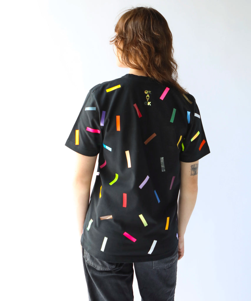 Woman wearing a black cotton t-shirt with colorful confetti sprinkles on it and a logo on the back saying 'OkayOk'