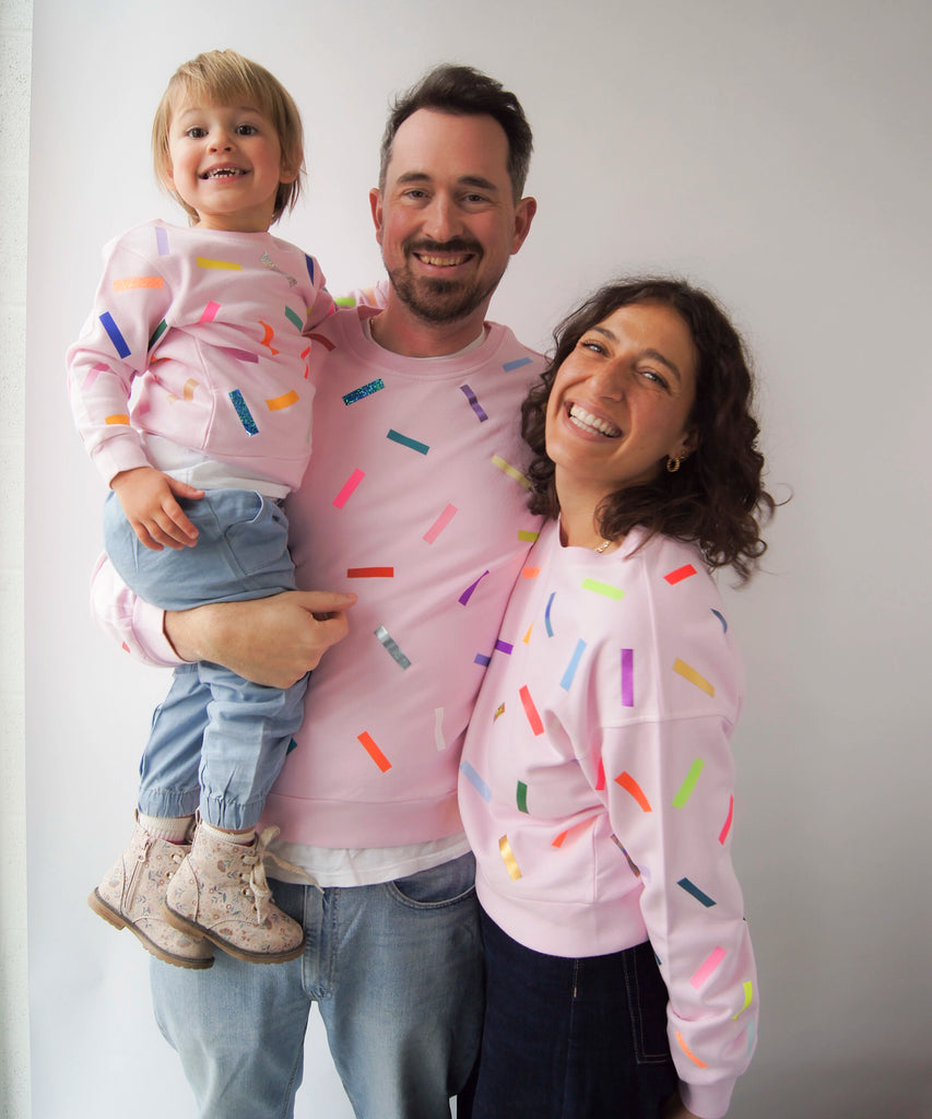 a woman, child and man wear matching cotton sweatshirt in pale pink with colourful confetti sprinkle graphic print