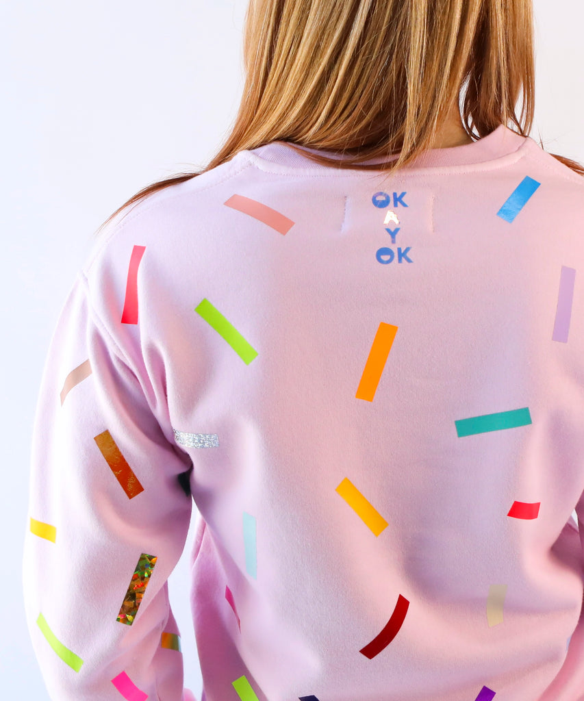 a close up of a woman wearing a pink cotton sweatshirt with colorful sprinkles confetti pattern and the branding 'okayok' on it