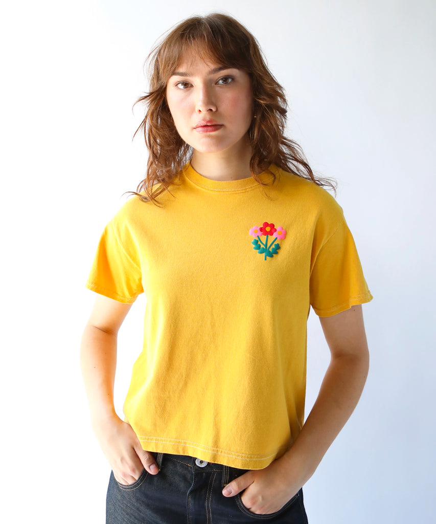 woman wearing a cropped, boxy fitting mustard yellow cotton t-shirt with a flower graphic on the left breast in pink and red