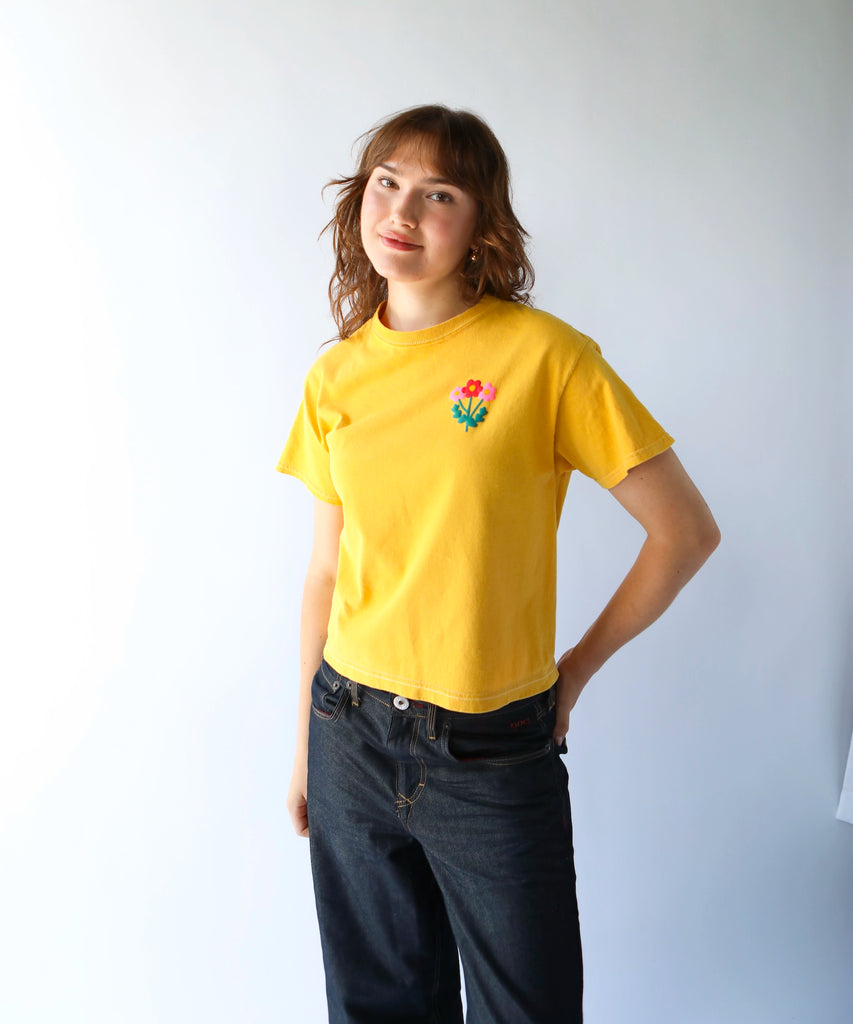 woman wearing a cropped, boxy fitting mustard yellow cotton t-shirt with a flower graphic on the left breast in pink and red