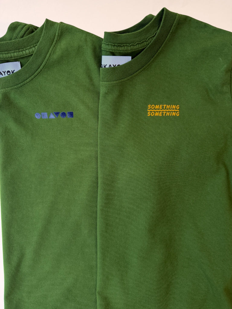 A moss green 100% cotton tshirt with two add-on graphic options: (left) with the purple 'okayok' Bauhaus shape logo, (right) a little 'something something' graphic with a squiggle line