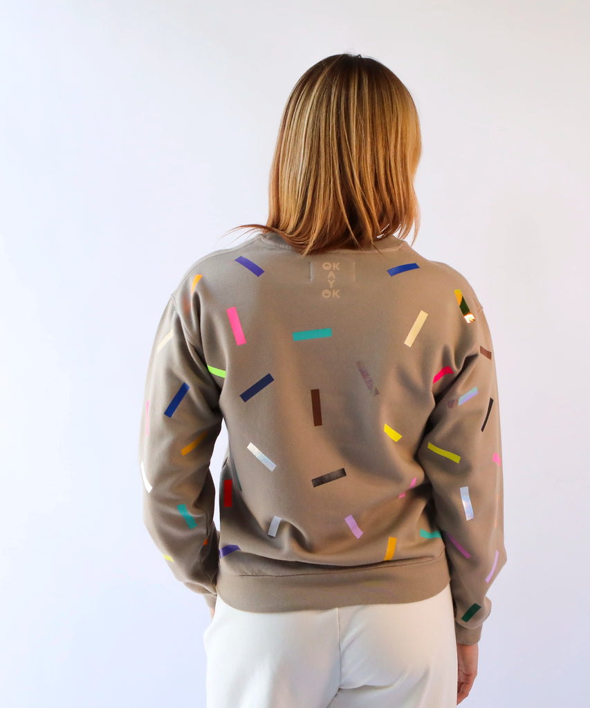 a woman wearing a taupe brown cotton sweatshirt with colorful sprinkles and the 'okayok' logo on the back