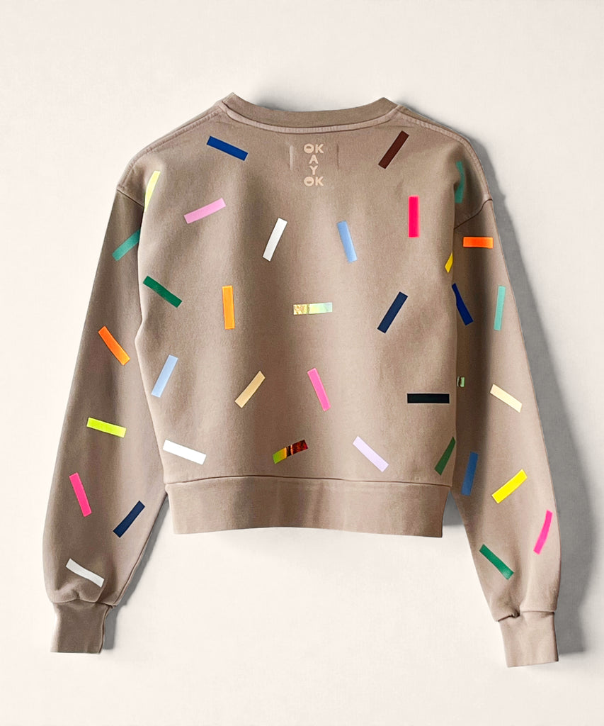A 100% cotton sweatshirt in taupe with colourful confetti sprinkle graphic print nd the 'okayok' logo on the back