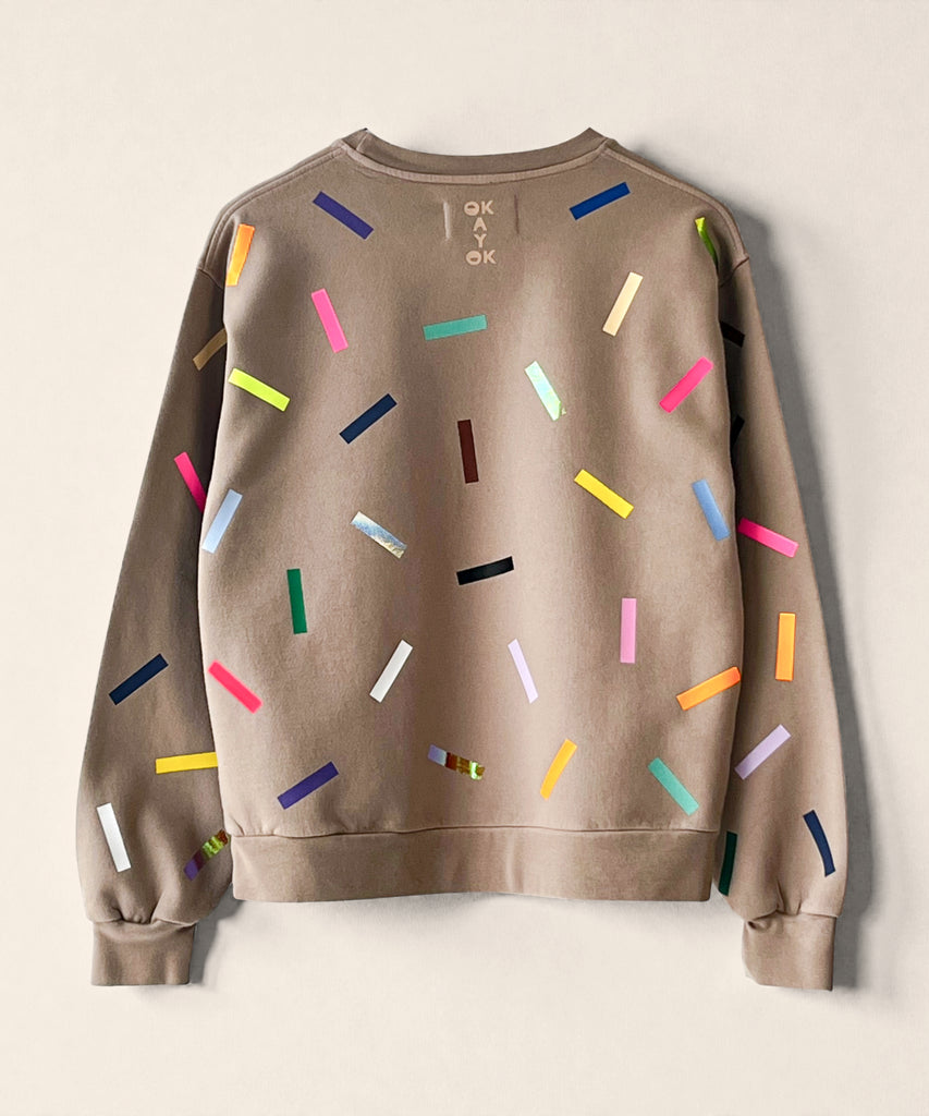 A 100% cotton sweatshirt in taupe with colourful confetti sprinkle graphic print and the 'okayok' logo on the back