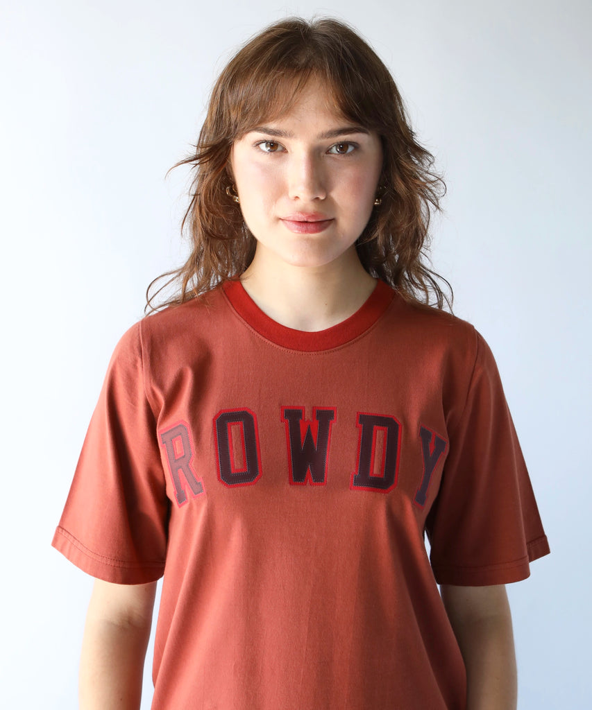 Person wearing a rust coloured 'ROWDY' cotton tshirt written with collegiate letters