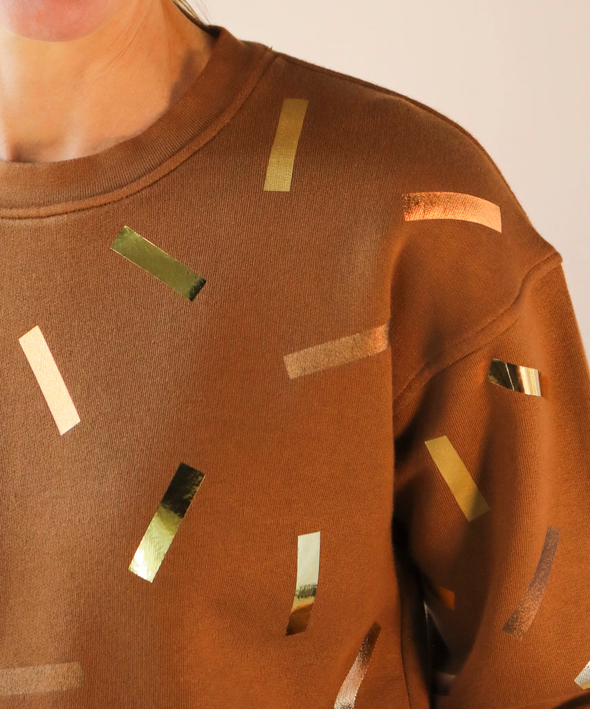 a woman wears a brown sweatshirt with gold and bronze confetti sprinkle print design