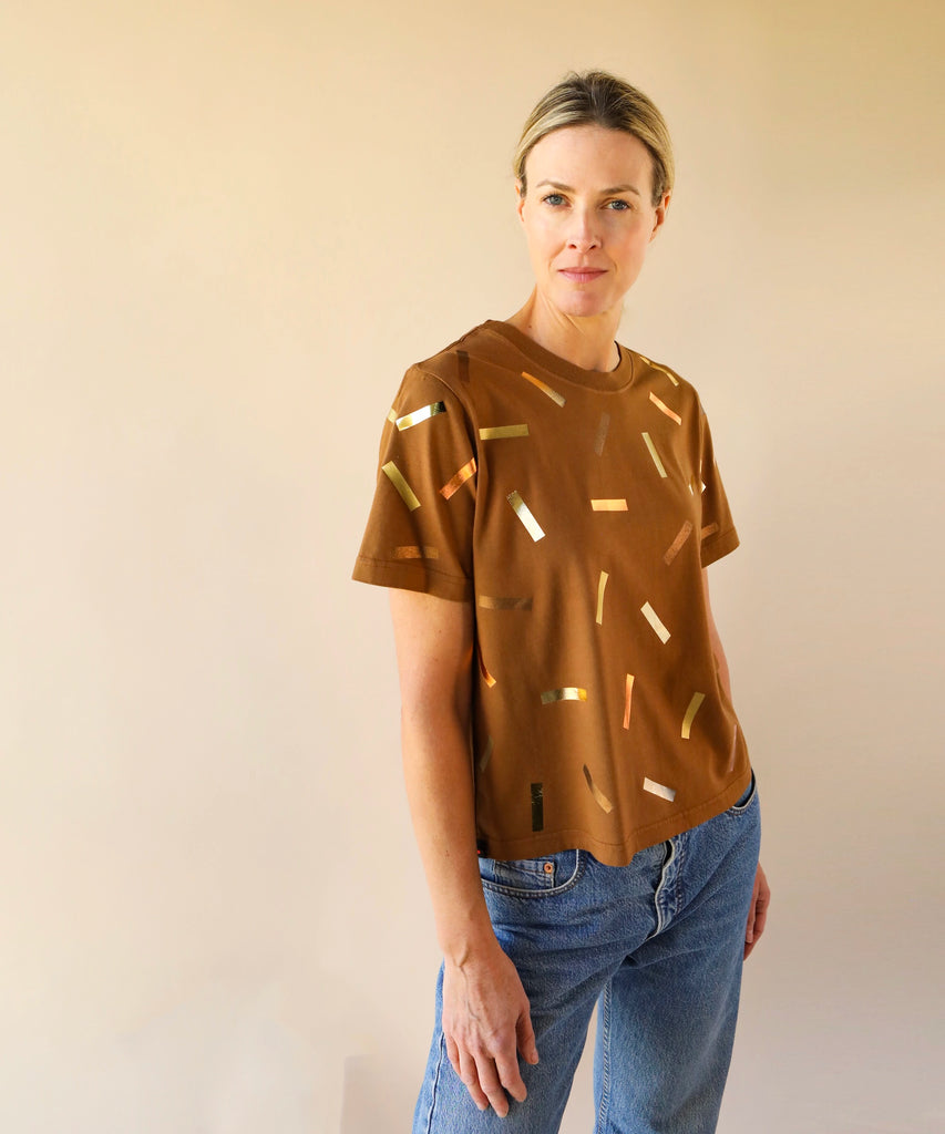 a woman wears a boxy cotton tshirt in tobacco brown with gold confetti sprinkle graphic print