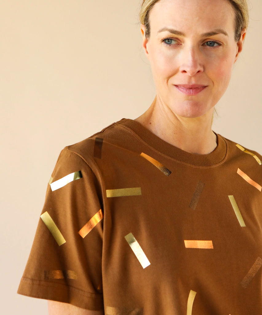 a woman wears a boxy cotton tshirt in tobacco brown with gold confetti sprinkle graphic print