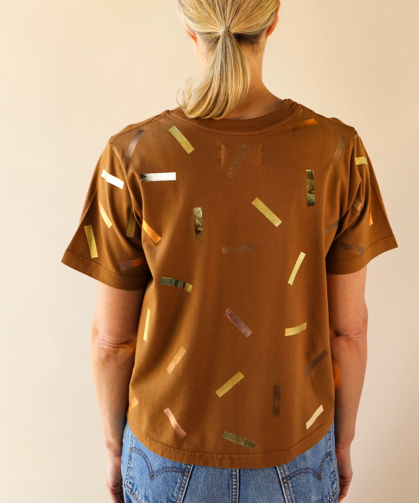 a woman wears a boxy cotton tshirt in tobacco brown with gold confetti sprinkle graphic print