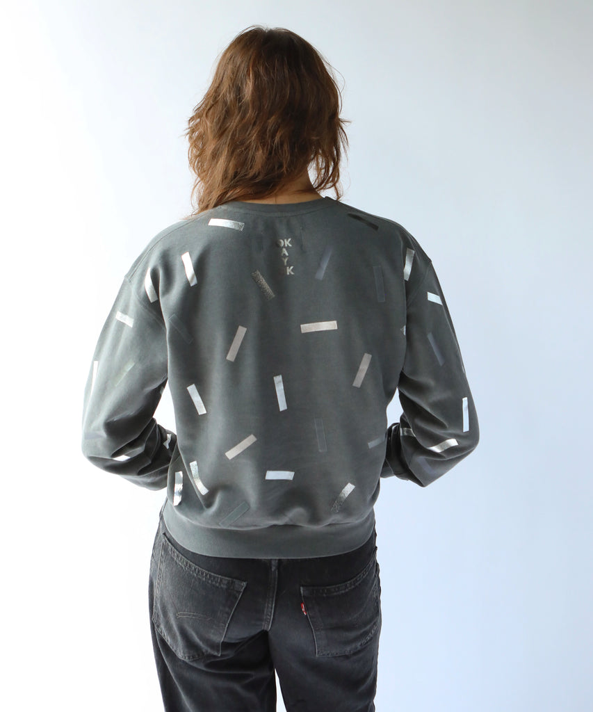 the back of a woman wearing a cotton sweatshirt in grey with silver confetti sprinkle graphic print with the words okayok on the back